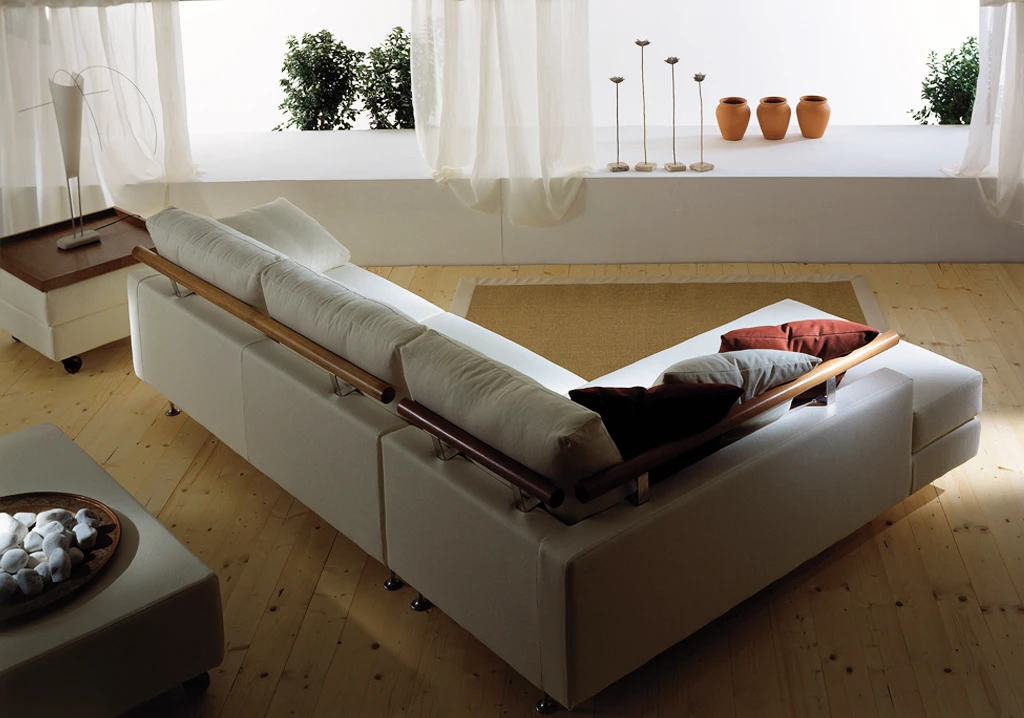 Vista posteriore di un divano bianco con chaise longue, con supporti schienali in legno, cuscini morbidi e ambiente minimalista con dettagli decorativi